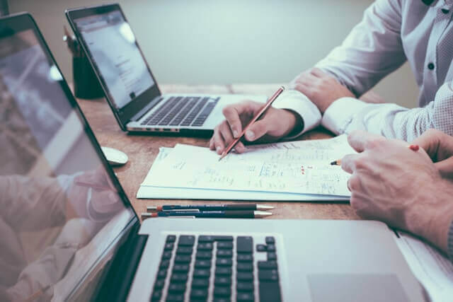 Zwei Personen vergleichen Unterlagen und Angebote an einem Tisch mit zwei Laptops